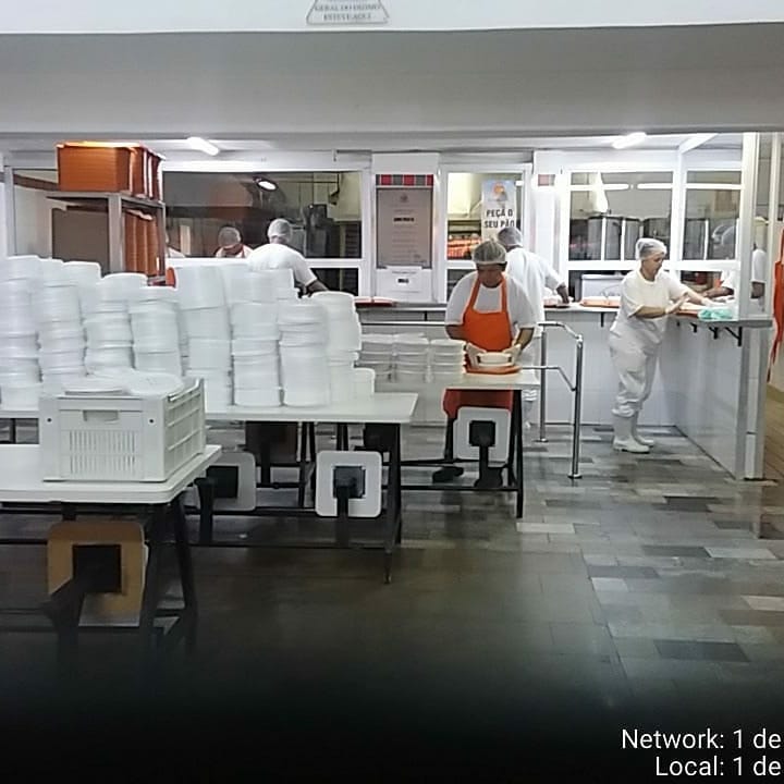 funcionários trabalham na cozinha de um restaurante, com roupas brancas e touca, ao lado de pacotes de marmitex de isopor. 